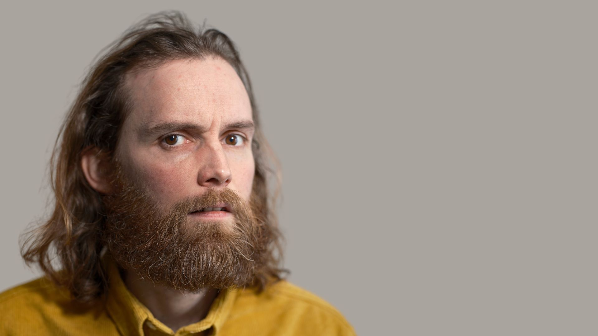 A man with long hair and a beard wearing a yellow shirt stares at the camera with a neutral expression