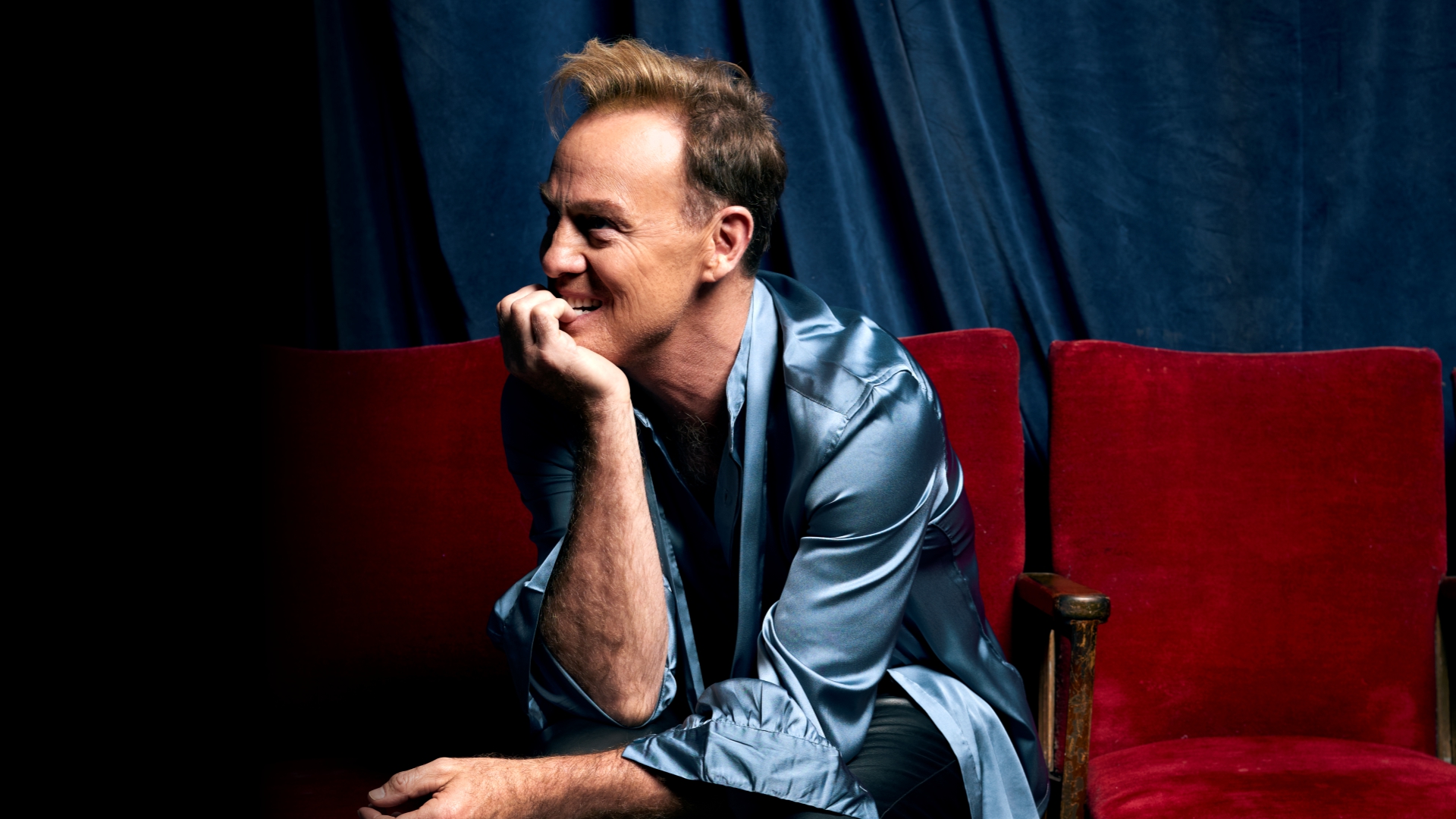 A man in a blue satin shirt sits ion a red sofa with his head in his right elbow on his knee and his head in his hand.