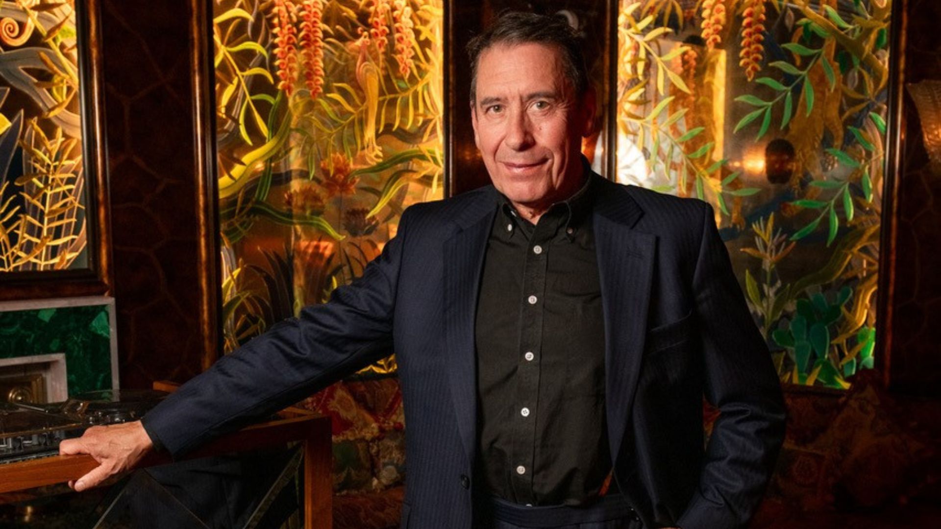Jools Holland in a black suit with colourful paintings of plants in the background