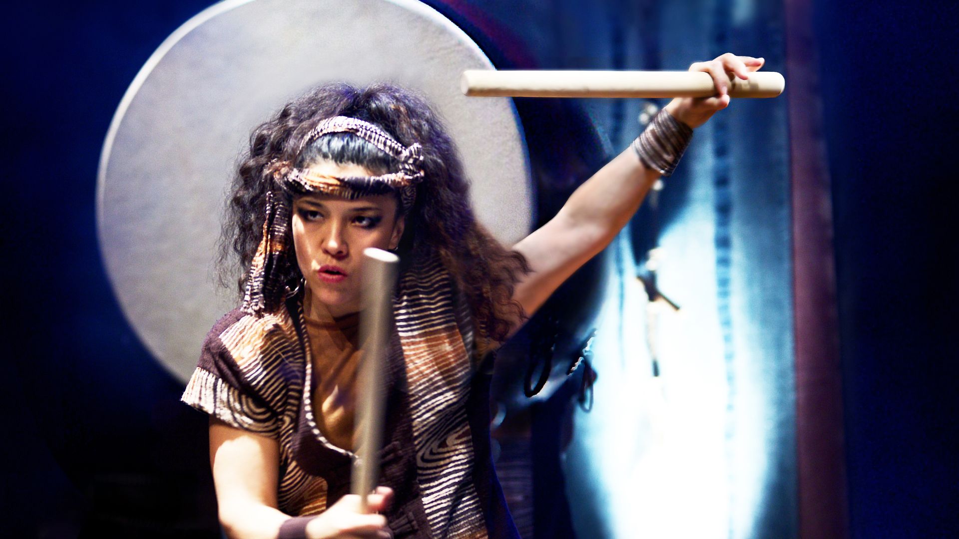 Woman in traditional Asian dress with drum sticks beating a drum.
